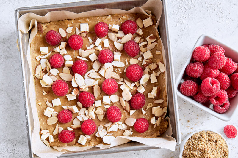 Unbaked blondies in parchment-lined baking pan topped with fresh raspberries, white chocolate, and sliced almonds