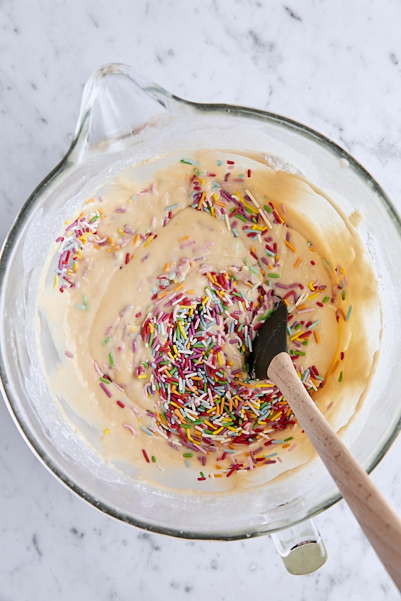 Rainbow sprinkles folded into cake batter in glass mixing bowl with spatula